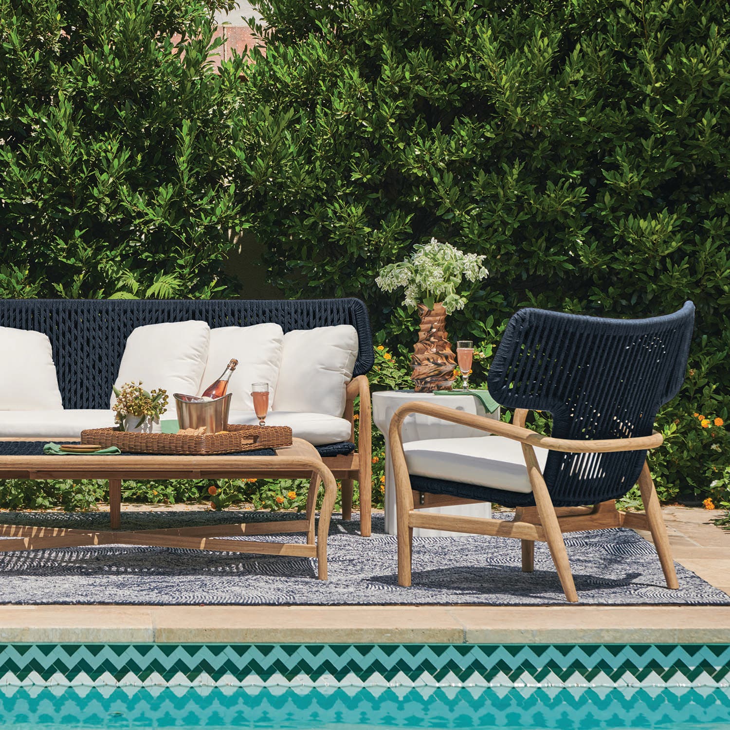 Poolside lounge grouping with a navy woven sofa and lounge chair, ivory cushions, and wood frames beside a pool with a lush green hedge behind