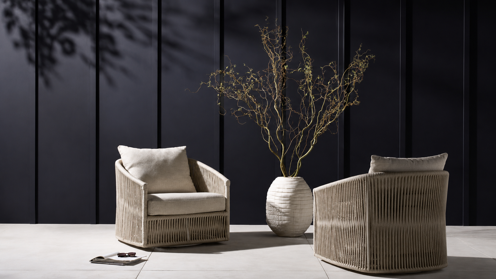 Two ivory woven lounge chairs arranged around a sculptural white vase with tall branches against a dark paneled wall