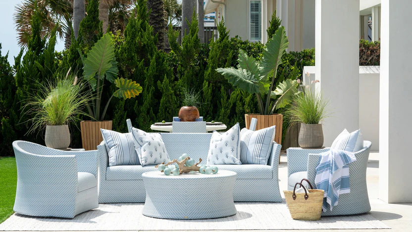 Coastal outdoor seating group with a light blue woven loveseat and swivel chairs around a round woven coffee table, framed by tropical greenery